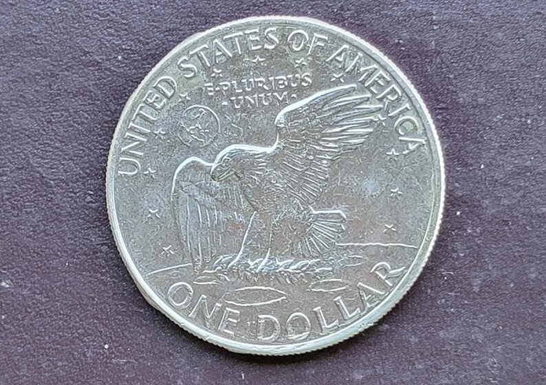 Op de afbeelding: Een zilveren dollar munt met een adelaar met uitgespreide vleugels, omringd door de woorden "UNITED STATES OF AMERICA" en "ONE DOLLAR". De munt heeft ook de inscriptie "E PLURIBUS UNUM".