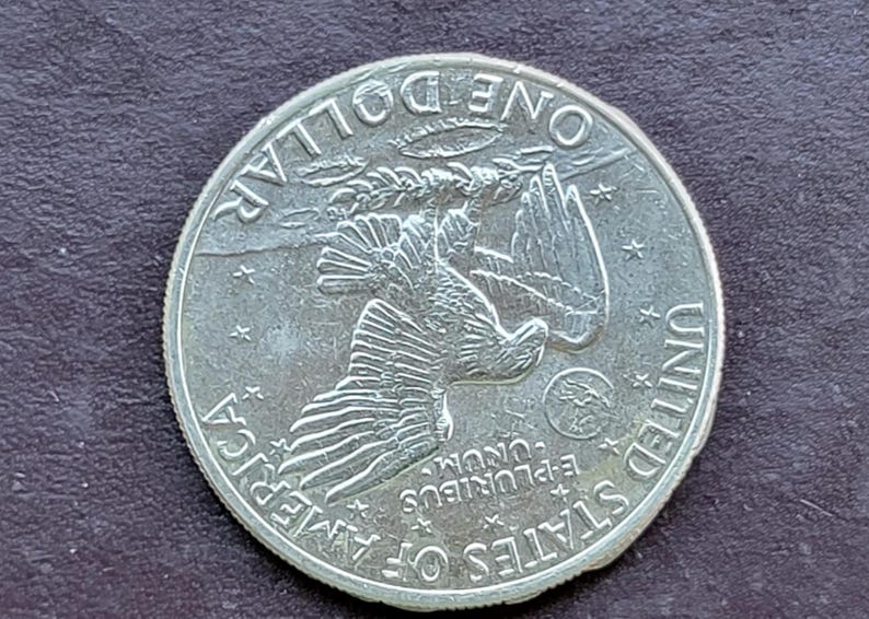 Op de afbeelding: Een zilveren dollar munt met een adelaar met uitgespreide vleugels, omringd door de woorden "ONE DOLLAR", "UNITED STATES OF AMERICA" en "E PLURIBUS UNUM". De munt heeft een gedetailleerd ontwerp en een licht versleten uiterlijk.