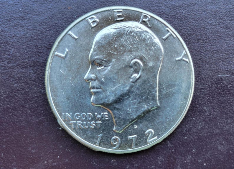 Op de afbeelding: Een zilveren Eisenhower dollar munt uit 1972. De munt toont een profiel van een man, het woord "LIBERTY" bovenaan, en "IN GOD WE TRUST" en de datum "1972" onderaan. De munt ligt op een donkere achtergrond.