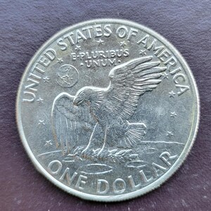 Op de afbeelding: Een zilveren &eacute;&eacute;n-dollar munt met een adelaarontwerp, met de inscripties "UNITED STATES OF AMERICA" en "ONE DOLLAR". De munt bevat ook de Latijnse zin "E PLURIBUS UNUM", wat "Uit velen, &eacute;&eacute;n" betekent.