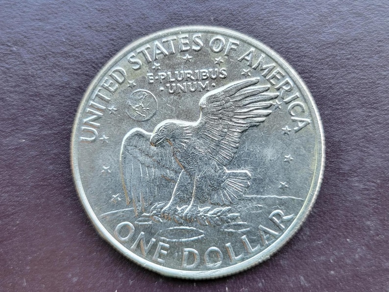 Op de afbeelding: Een zilveren &eacute;&eacute;n-dollar munt met een adelaarontwerp, met de inscripties "UNITED STATES OF AMERICA" en "ONE DOLLAR". De munt bevat ook de Latijnse zin "E PLURIBUS UNUM", wat "Uit velen, &eacute;&eacute;n" betekent.