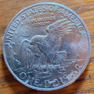 Op de afbeelding: Een zilveren dollar munt met de woorden "UNITED STATES OF AMERICA" en "ONE DOLLAR" in reli&euml;f langs de randen. De munt toont een adelaar met uitgespreide vleugels en de inscriptie "E PLURIBUS UNUM".