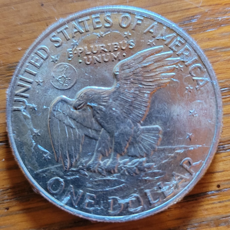 Op de afbeelding: Een zilveren dollar munt met de woorden "UNITED STATES OF AMERICA" en "ONE DOLLAR" in reli&euml;f langs de randen. De munt toont een adelaar met uitgespreide vleugels en de inscriptie "E PLURIBUS UNUM".