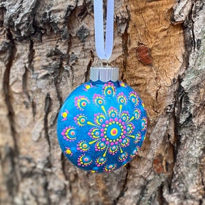 May include: A blue glass Christmas ornament with a hand-painted design. The ornament features a central circular pattern with pink, yellow, and blue dots. A white ribbon is attached for hanging. The ornament is approximately 3 inches in diameter.