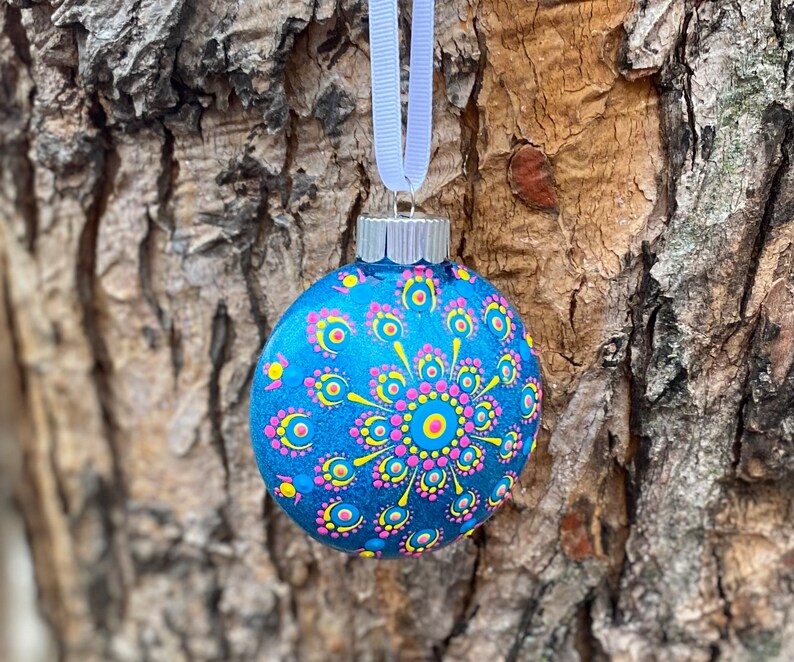 May include: A blue glass Christmas ornament with a hand-painted design. The ornament features a central circular pattern with pink, yellow, and blue dots. A white ribbon is attached for hanging. The ornament is approximately 3 inches in diameter.