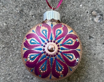 May include: A round glass ornament with a gold background and a pink and teal mandala design. The ornament has a silver top and a red ribbon.