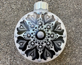 May include: A round, silver Christmas ornament with a black, gray, and silver floral design. The ornament has a silver cap and a black string for hanging. The design is symmetrical and detailed, with a central circle and radiating petal shapes.