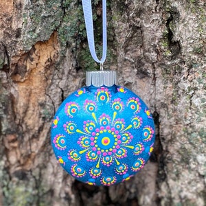 May include: A round, blue Christmas ornament with a hand-painted mandala design. The ornament features a central pink circle surrounded by yellow and pink dots. A white ribbon is attached for hanging.