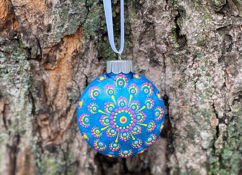 May include: A round, blue Christmas ornament with a hand-painted mandala design. The ornament features a central pink circle surrounded by yellow and pink dots. A white ribbon is attached for hanging.