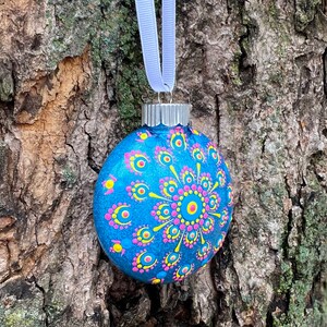 May include: A hand-painted blue glass Christmas ornament with a decorative mandala design. The ornament features a pattern of yellow, pink, and blue dots and lines. A silver metal cap and a light blue ribbon are attached for hanging.