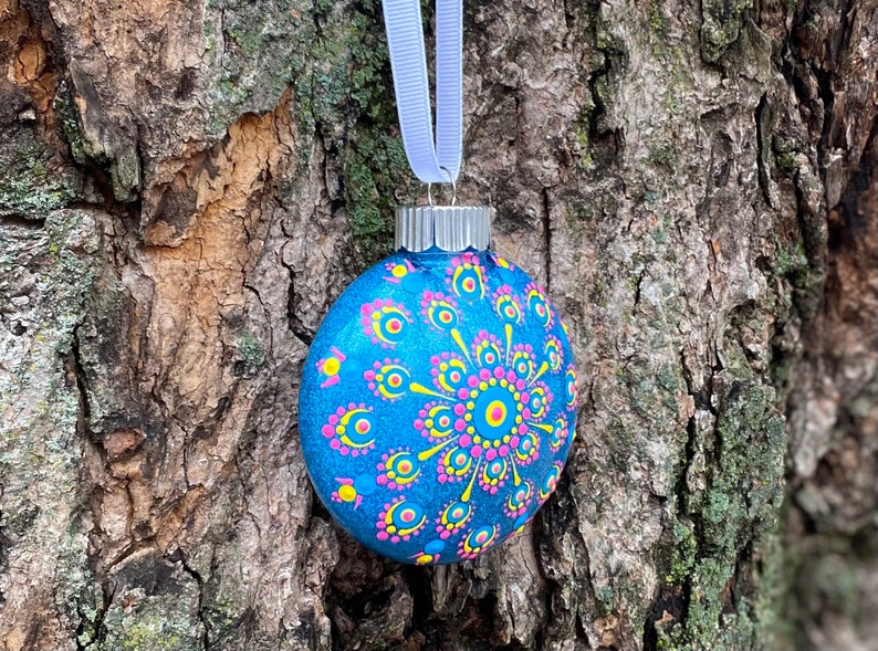 May include: A hand-painted blue glass Christmas ornament with a decorative mandala design. The ornament features a pattern of yellow, pink, and blue dots and lines. A silver metal cap and a light blue ribbon are attached for hanging.