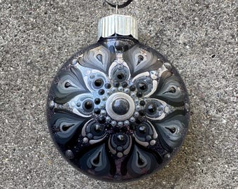 May include: A round, black Christmas ornament with a silver cap and black ribbon. The ornament is decorated with a symmetrical floral design in shades of gray, silver, and black, with a glittery finish.