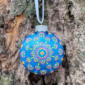 May include: A blue Christmas ornament with a hand-painted mandala design. The ornament features a central pink and yellow flower-like pattern, surrounded by smaller yellow and pink dots. It hangs from a white ribbon.