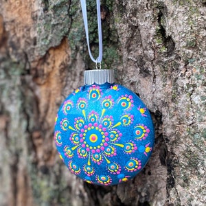 May include: A blue glass Christmas ornament with a silver cap and white ribbon. The ornament is decorated with a mandala design in pink, yellow, and blue dots. The ornament is hanging from a tree.