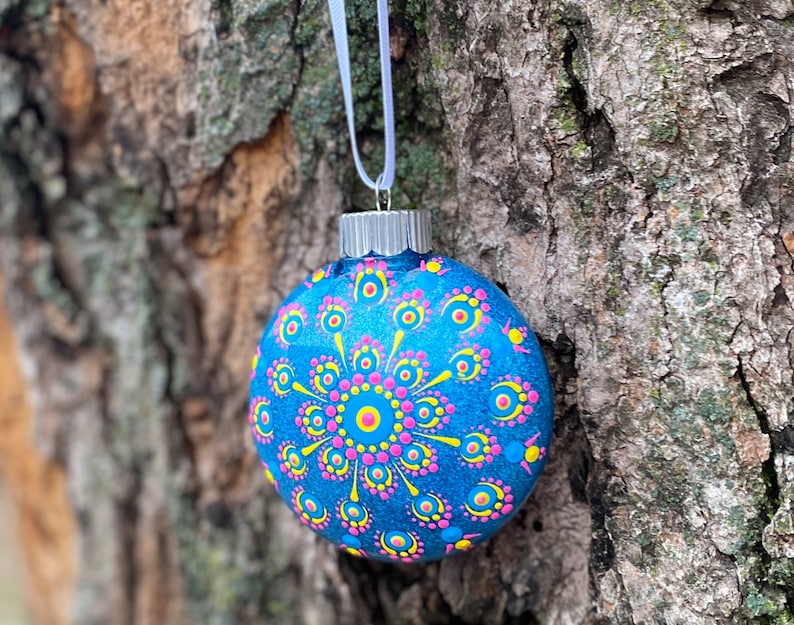 May include: A blue glass Christmas ornament with a silver cap and white ribbon. The ornament is decorated with a mandala design in pink, yellow, and blue dots. The ornament is hanging from a tree.