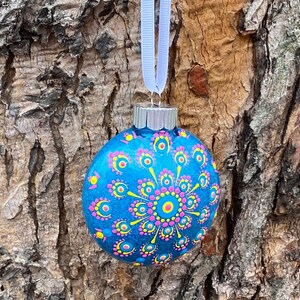 May include: A blue Christmas ornament with a hand-painted design. The ornament features a central circular pattern in pink and yellow, surrounded by smaller pink and yellow dots. A white ribbon is attached for hanging.