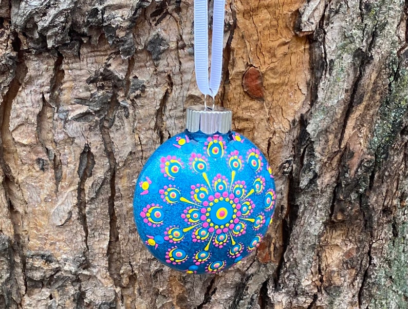 May include: A blue Christmas ornament with a hand-painted design. The ornament features a central circular pattern in pink and yellow, surrounded by smaller pink and yellow dots. A white ribbon is attached for hanging.