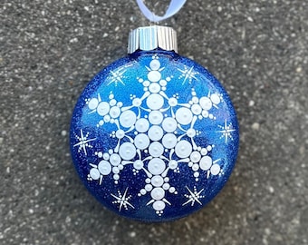 May include: A round, blue Christmas ornament with a white snowflake design. The ornament has a glittery finish and a silver cap with a white ribbon for hanging. The snowflake is made of white dots.