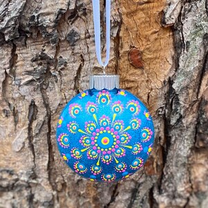 May include: A blue glass Christmas ornament with a hand-painted mandala design. The ornament features a central yellow circle surrounded by pink and yellow dots, with a white ribbon for hanging. The background is a textured tree trunk.