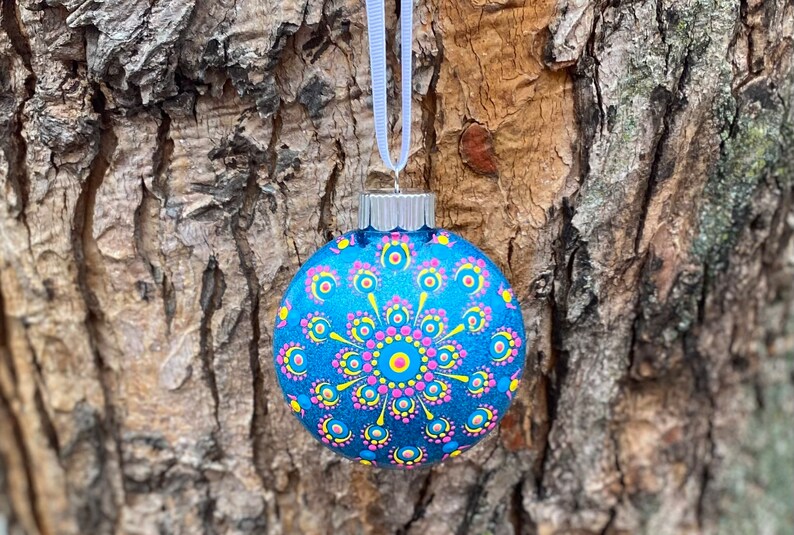 May include: A blue glass Christmas ornament with a hand-painted mandala design. The ornament features a central yellow circle surrounded by pink and yellow dots, with a white ribbon for hanging. The background is a textured tree trunk.