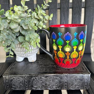 May include: A black ceramic mug with a vibrant, multi-colored dot art pattern featuring concentric circles in shades of red, orange, yellow, green, blue, and purple. The interior of the mug is a solid, bright red, and it has a curved black handle.