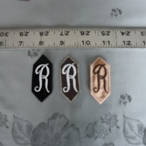 May include: Three fabric patches with the letter "R" embroidered in white, brown, and black thread. The patches are in the shape of a hexagon.