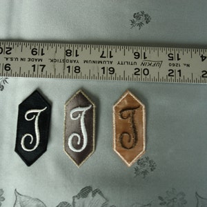 以下が含まれることがあります： 六角形の刺繍ワッペン3枚。白い文字「J」が黒地に、茶色地に、ベージュ地に刺繍されている。