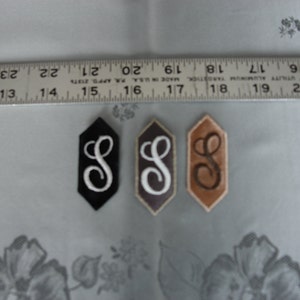 May include: Three personalised name tags in black, brown, and tan, each with a white script letter "S". The tags are in an elongated hexagon shape. A metal ruler is in the background. The ruler measures up to 25 inches.