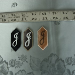 Puede incluir: Tres parches de tela con la letra "J" en una fuente cursiva. Los parches son negros, marrones y beige.