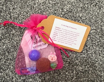 May include: A small pink mesh bag with a collection of trinkets, including a purple marble, a brown coin, and a small bunny-shaped charm. A tag reads "A Little Bag of Happiness" with a list of items and their meanings.