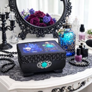 Könnte beinhalten: Schwarze dekorative Box mit türkisfarbenem Akzent und einem Totenkopf- und Trank-Design auf dem Deckel. Die Box steht auf einem weißen Schminktisch mit einem schwarzen Spitzentischläufer, einer blauen Glasflasche und einem schwarzen Spiegel.