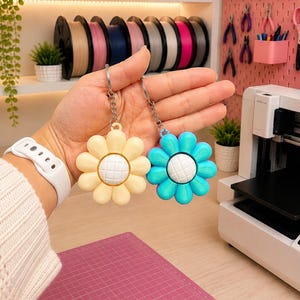 May include: Two flower-shaped keychains, one in pale yellow and one in bright blue, each featuring a white center with a grid pattern. The keychains are attached to silver-toned metal split rings and chains, held up to display the floral design.