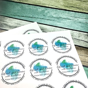 Op de afbeelding: Vellen ronde stickers met de tekst "Refresh and Renew with eucalyptus YOGA MAT SPRAY." Het ontwerp bevat een aquarel-achtige blauwe en groene splash. De stickers liggen op een houten achtergrond.