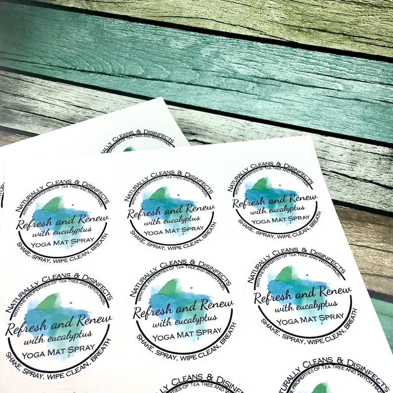 Op de afbeelding: Vellen ronde stickers met de tekst "Refresh and Renew with eucalyptus YOGA MAT SPRAY." Het ontwerp bevat een aquarel-achtige blauwe en groene splash. De stickers liggen op een houten achtergrond.