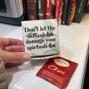 May include: A white box with green leaves and the text "Don't let the difficul-tea damage your spirtuali-tea" on it. A red tea bag with the text "TWININGS Chai" and the words "Fine black tea perfectly balanced with the sweet and savoury spice flavours of cinnamon, cardamom, cloves and ginger. 1 tea bag" on it.