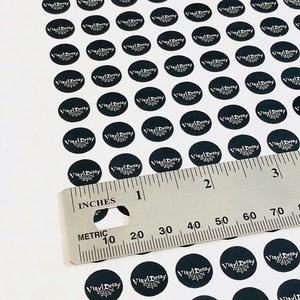 May include: A white surface with a ruler and many black round stickers with the text "Vinyl Decay" in white lettering. The stickers are arranged in rows and columns.