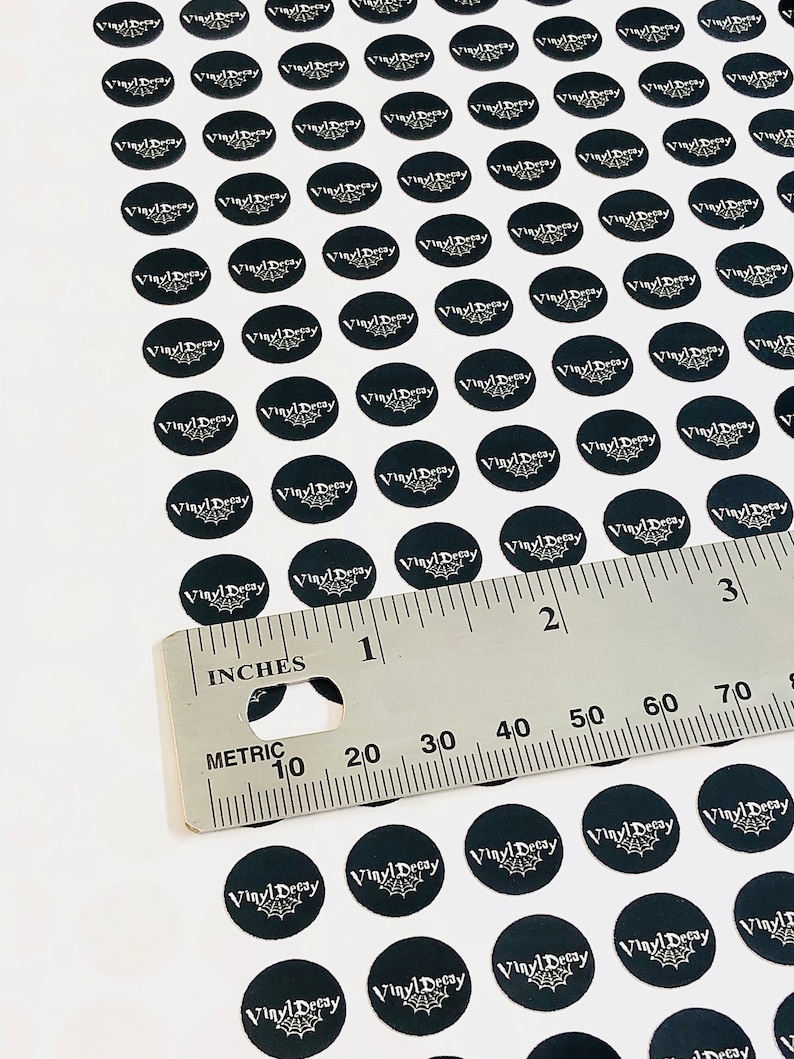 May include: A white surface with a ruler and many black round stickers with the text "Vinyl Decay" in white lettering. The stickers are arranged in rows and columns.
