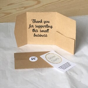 K&ouml;nnte beinhalten: Eine braune Papier-Dankeskarte mit dem Text "Vielen Dank f&uuml;r die Unterst&uuml;tzung dieses kleinen Unternehmens". Die Karte ist in der Mitte gefaltet und hat eine kleine Tasche auf der Vorderseite. Ein kleiner brauner Umschlag mit einem wei&szlig;en Kreis mit den Buchstaben "SC" in Schwarz ist im Hintergrund zu sehen.