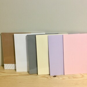 May include: Six sheets of cardstock in different colours: brown, white, grey, yellow, purple, and pink. The sheets are arranged in a row on a wooden surface.