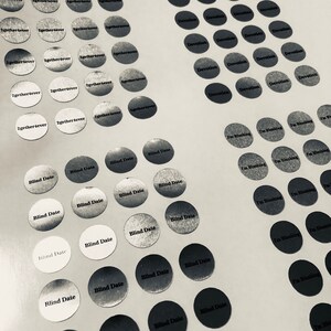 May include: A close-up of a variety of round stickers with different phrases printed on them. The stickers are silver and have black text. The phrases include "Blind Date", "I'm Blushing", and "2gether4ever".