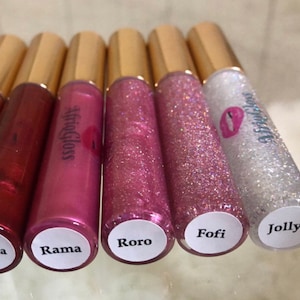 May include: Six clear lip gloss tubes with gold caps and white labels. The labels have the names "Rama", "Roro", "Fofi", "Jolly", and "#afrigloss" written on them.