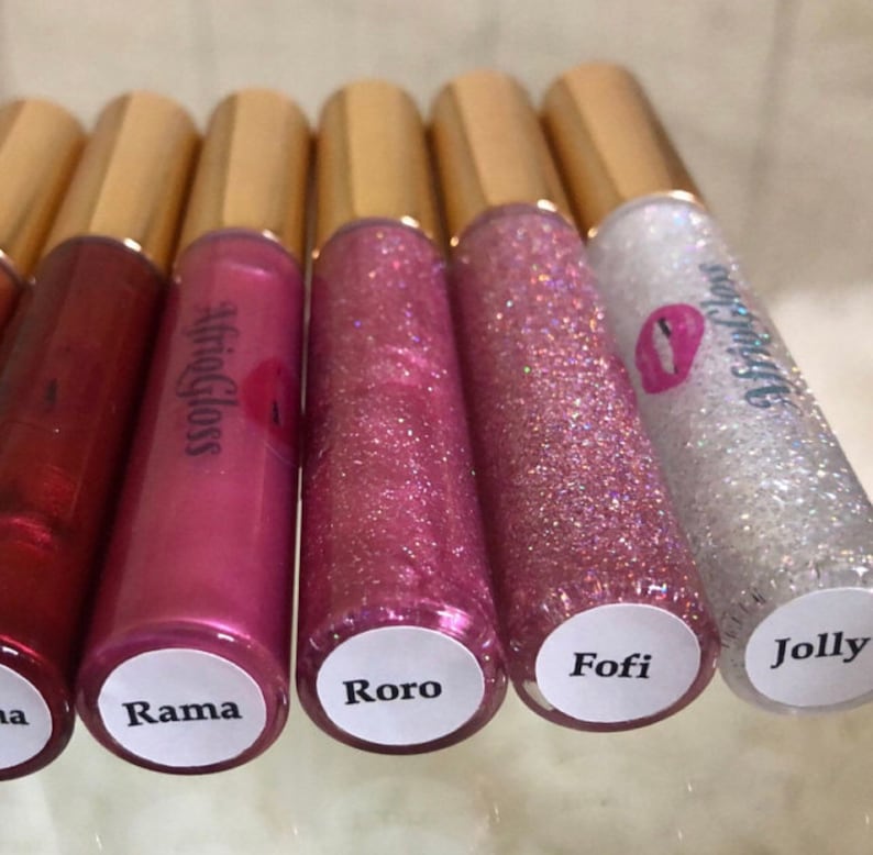 May include: Six clear lip gloss tubes with gold caps and white labels. The labels have the names "Rama", "Roro", "Fofi", "Jolly", and "#afrigloss" written on them.