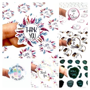 Op de afbeelding: Een verzameling decoratieve stickers met verschillende ontwerpen. Sommige stickers zeggen "Thank You" omringd door een veer- en bloemenkrans. Andere stickers zeggen "Let it Stain" en "Mail Time".