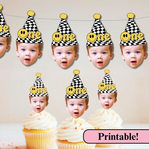 Op de afbeelding: Een banner met lachende gezichten met zwart-wit geblokte feesthoeden met het woord "One" en een smiley. Cupcakes met witte glazuur staan eronder. Het woord "Printable!" staat in een roze banner.