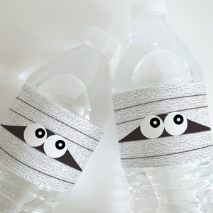 May include: Two clear plastic water bottles with white and gray mummy-themed labels. The labels have black eyes and a black triangle shape.