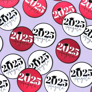 Pode incluir: Autocolantes circulares com o tema da formatura com o texto "2025 Angela" em preto e branco, e vermelho e branco. Uma imagem de um chap&eacute;u de formatura est&aacute; inclu&iacute;da. Os autocolantes est&atilde;o espalhados num fundo lil&aacute;s claro.
