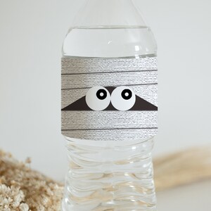 May include: A clear plastic water bottle with a white label that has a mummy design with two eyes peeking out from under the bandages. The label has a wood grain texture.