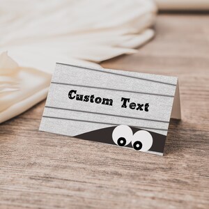 May include: A white card with a woodgrain texture and the text "Custom Text" printed in black. Two white eyes peek out from behind a brown shape at the bottom of the card.