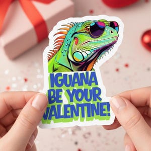 Puede incluir: Una colorida pegatina con un iguana de dibujos animados con gafas de sol. El iguana es verde, azul y naranja. La pegatina tiene el texto "IGUANA BE YOUR VALENTINE!" en azul y verde. La pegatina está sujeta entre dos manos.