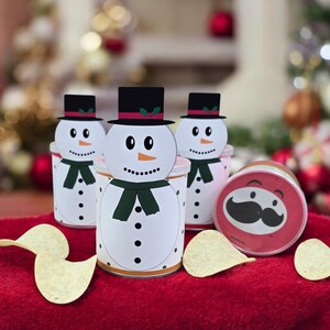 Könnte beinhalten: Drei Schneemann-förmige Behälter mit schwarzen Zylindern und grünen Schals sowie ein roter Behälter mit einem schwarzen Schnurrbart. Die Behälter stehen auf einer roten Oberfläche mit zwei Kartoffelchips.